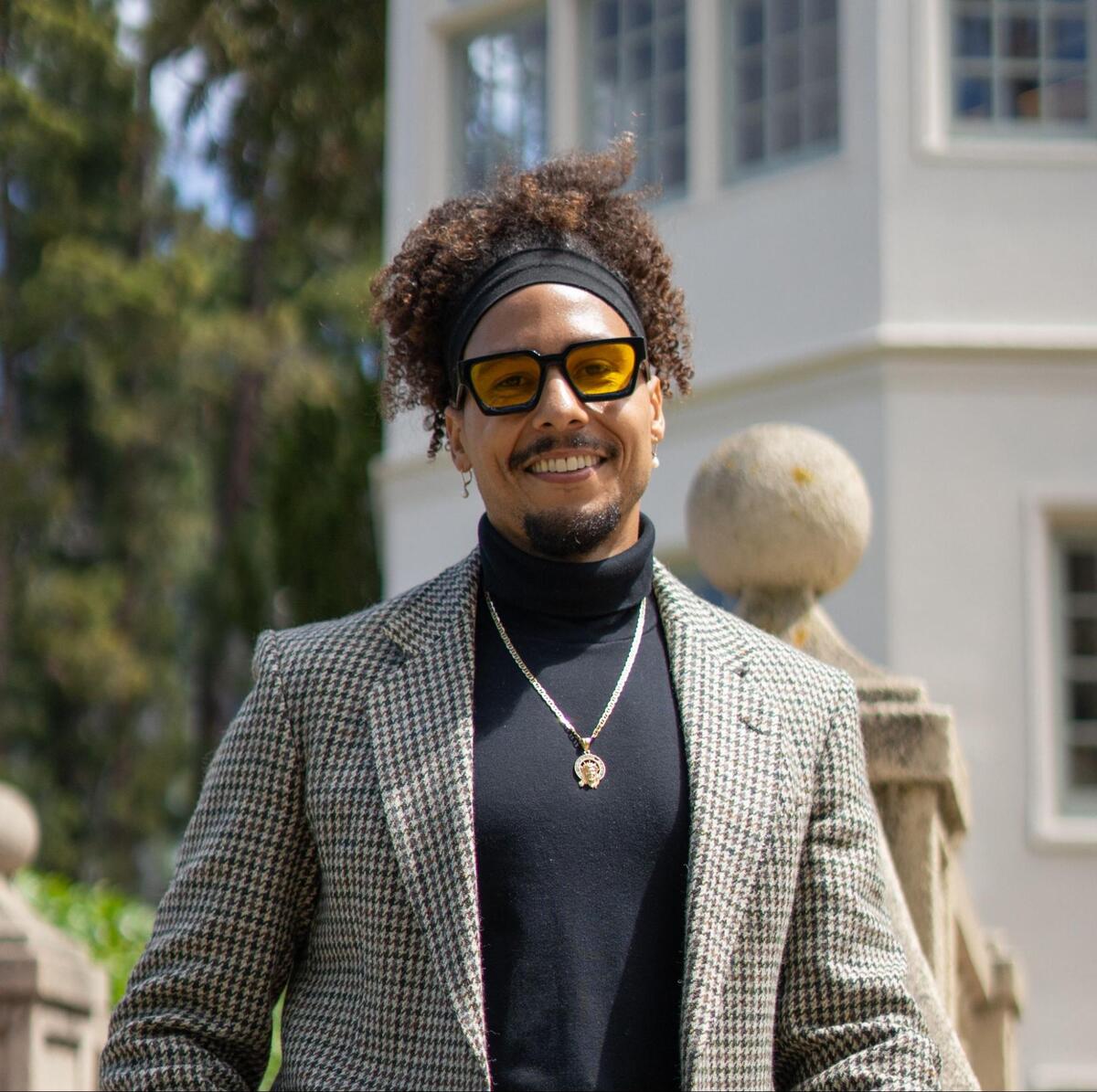 A picture of Daniel Lobo wearing yellow sunglasses, a black turtleneck shirt, and a gingham blazer.
