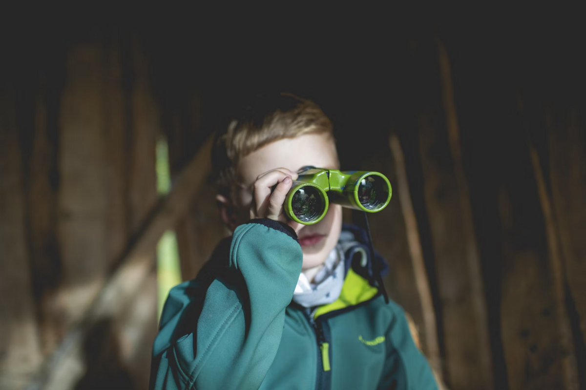 A kid in a teal jacket looking through a pair of green binoculars