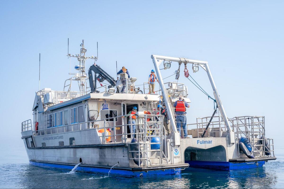 Research boat with A-frame on the boat deck to lower fiber optic cable into the water. Multiple researchers on board with orange life vests and blue hard hats.