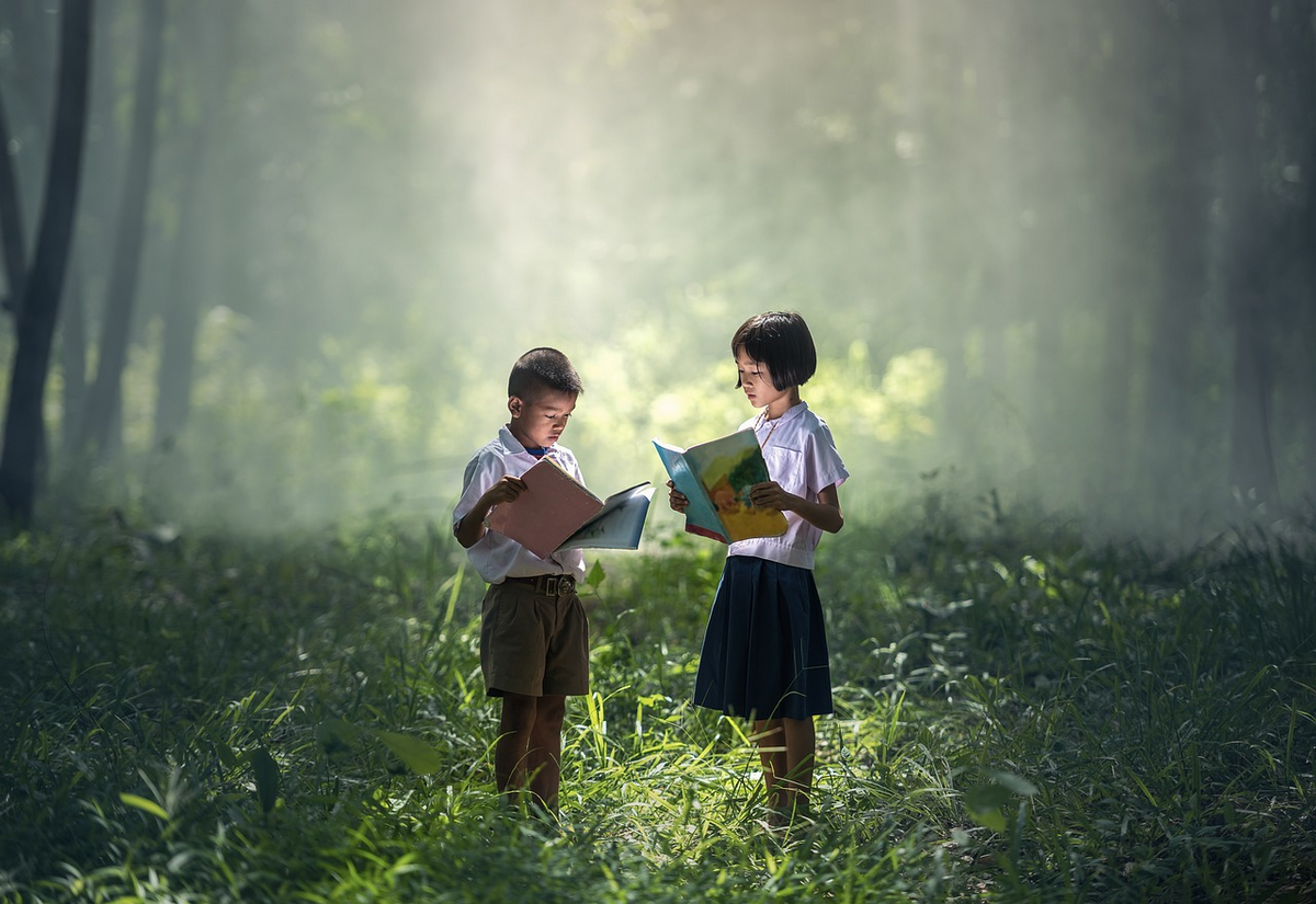 2 children of Asian descent, 1 boy and 1 girl, stand in a clearing in the forest reading books