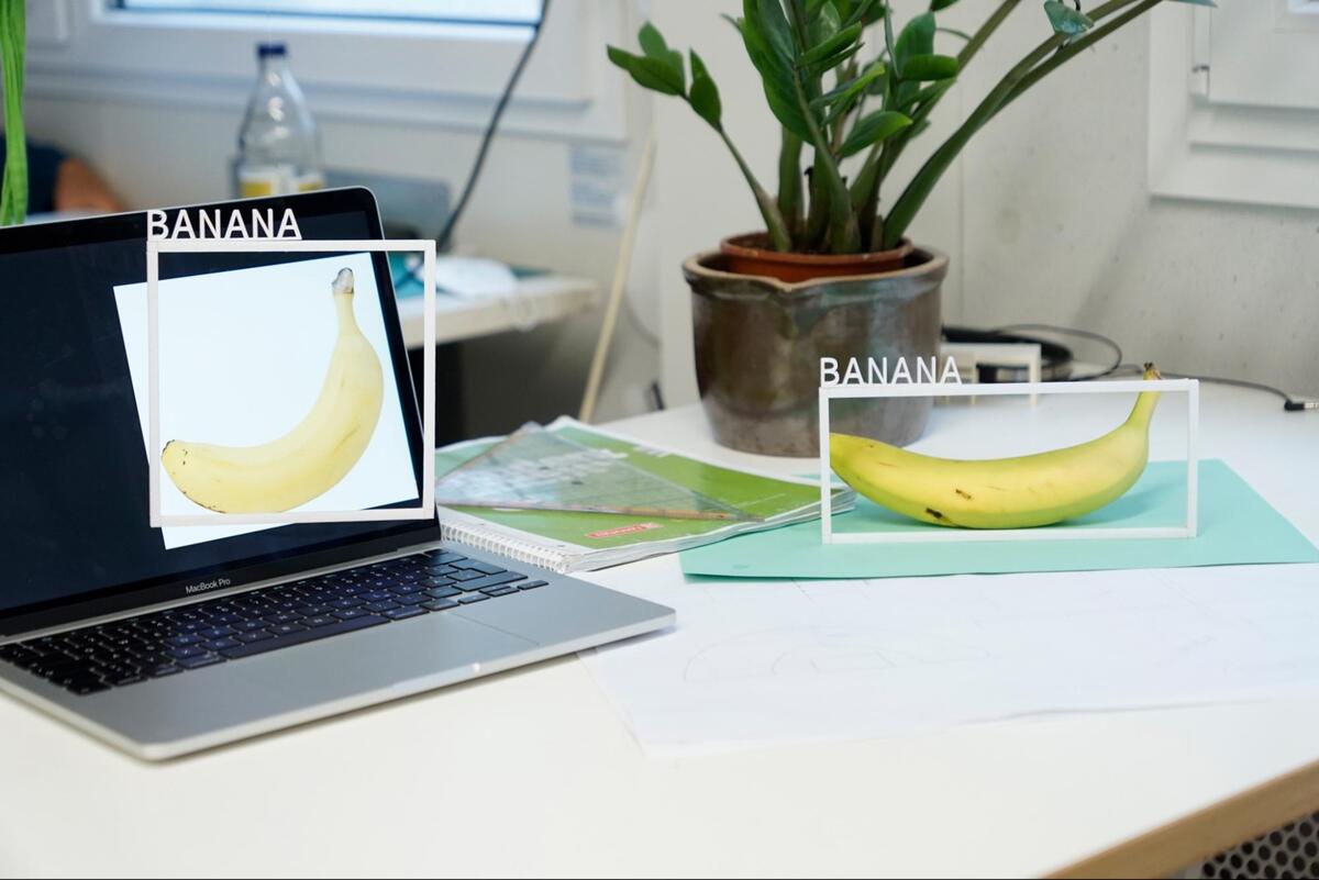 A real banana sitting next to a picture of a banana on a computer, both have a white box around them with the label "Banana".
