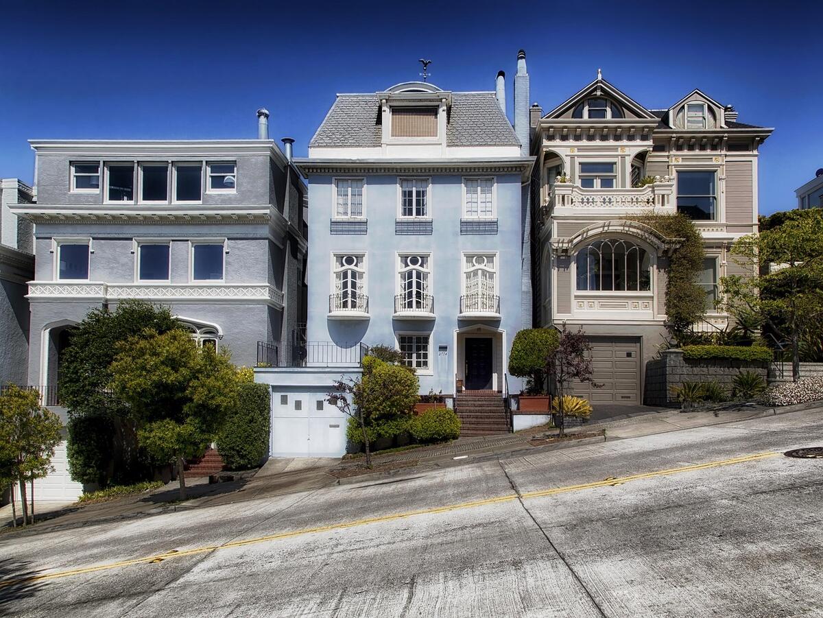 A picture of the front of 3 multi-story houses close together on a steep street. From left to right, the houses are grey, light blue, and tan.