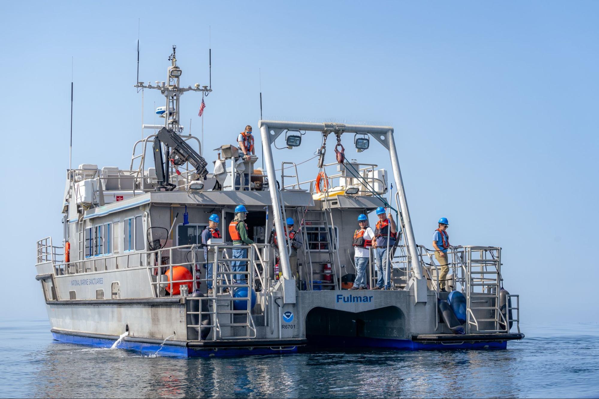 Research boat with A-frame on the boat deck to lower cable into the water. Multiple researchers on board with orange life vests and blue hard hats.